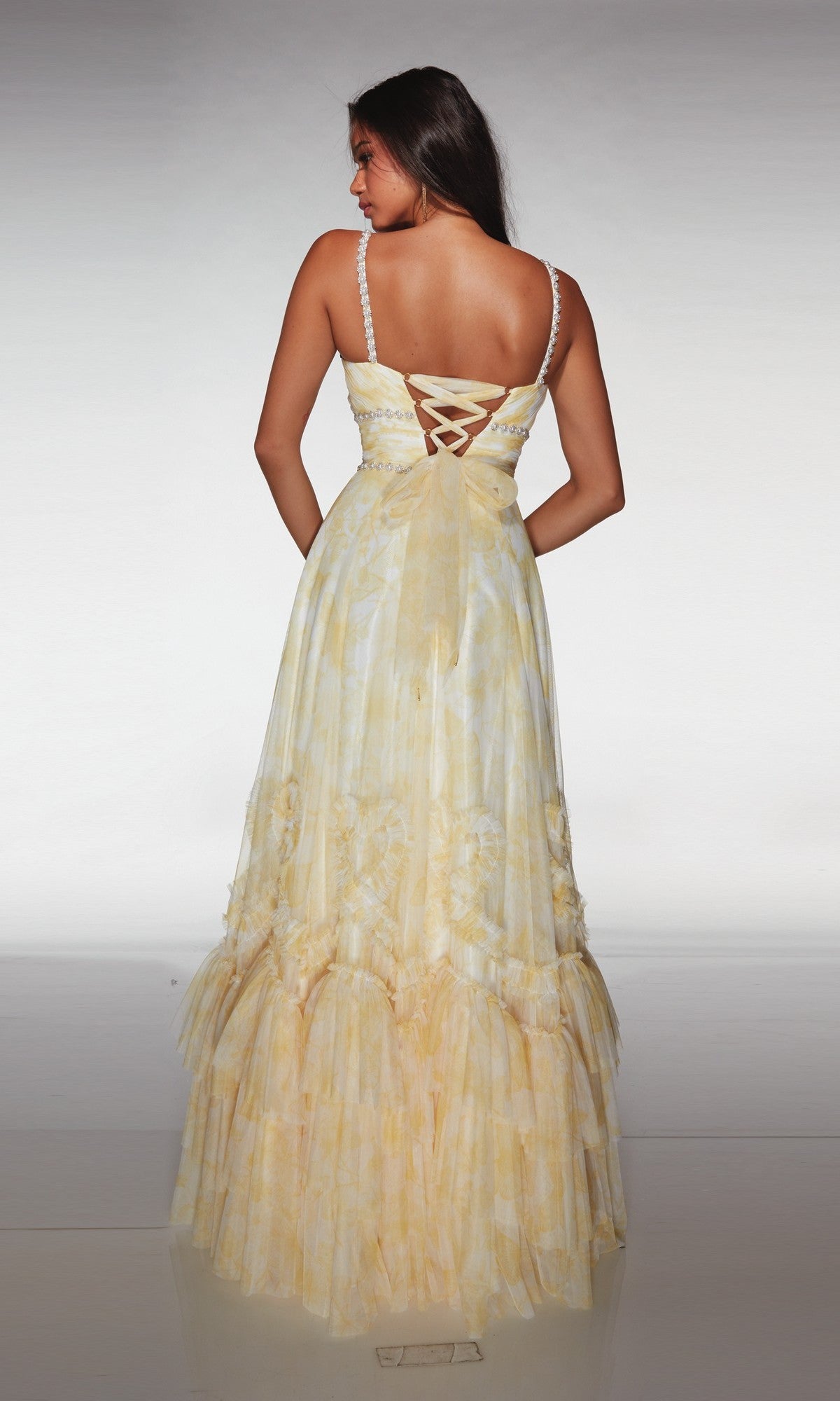A woman stands with her back to the camera, showcasing the Alyce 62063—a long, light yellow floral formal dress with thin straps and a lace-up back—against a plain background.
